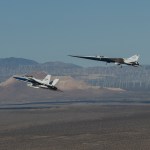 NASA’s X-59 flies above the Mojave Desert with a NASA F-15 off its left wing. The X-59’s landing gear is down and below it the desert landscape stretches toward rows of windmills in front of mountains.