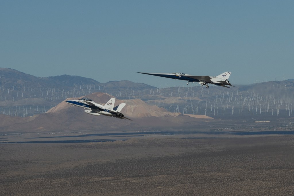NASA’s X-59 flies above the Mojave Desert with a NASA F-15 off its left wing. The X-59’s landing gear is down and below it the desert landscape stretches toward rows of windmills in front of mountains.