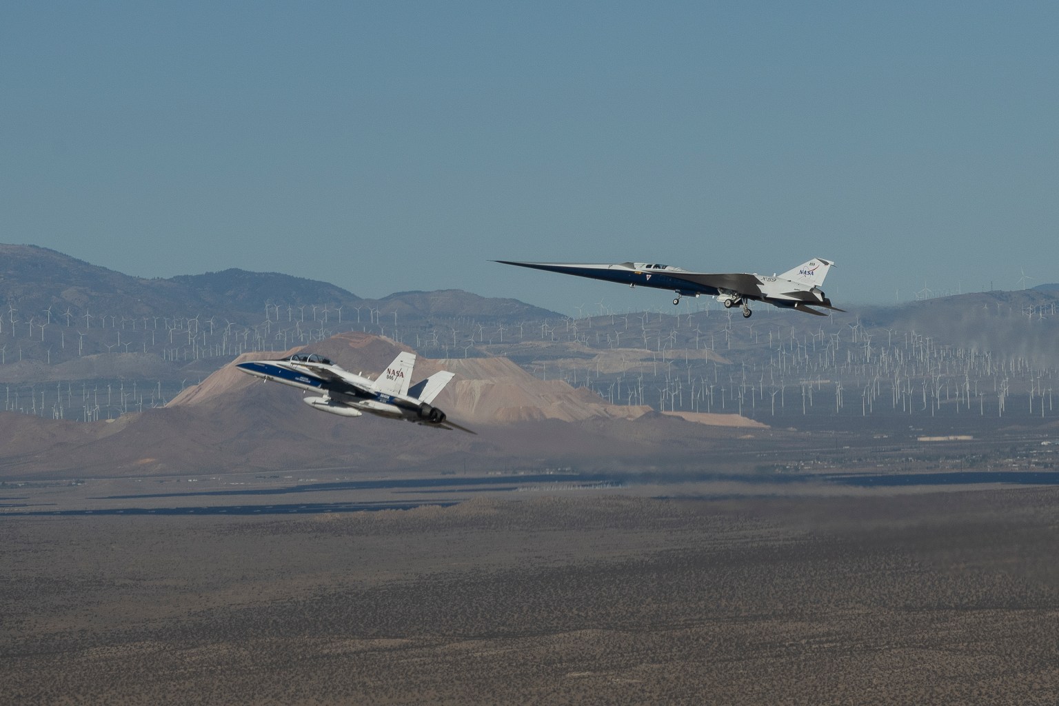 NASA’s X-59 flies above the Mojave Desert with a NASA F-15 off its left wing. The X-59’s landing gear is down and below it the desert landscape stretches toward rows of windmills in front of mountains.