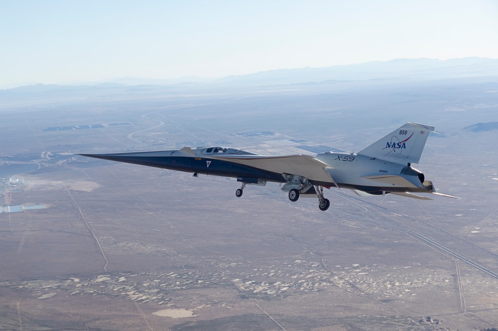 NASA’s X-59 is seen in flight, with a blue sky and mountains behind it and land below it. The aircraft’s long nose and distinct silhouette are visible as it ferries to NASA’s Armstrong Flight Research Center in Edwards, California.