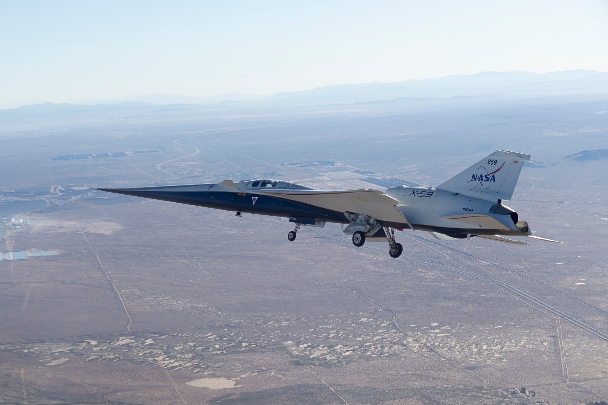 NASA’s X-59 is seen in flight, with a blue sky and mountains behind it and land below it. The aircraft’s long nose and distinct silhouette are visible as it ferries to NASA’s Armstrong Flight Research Center in Edwards, California.