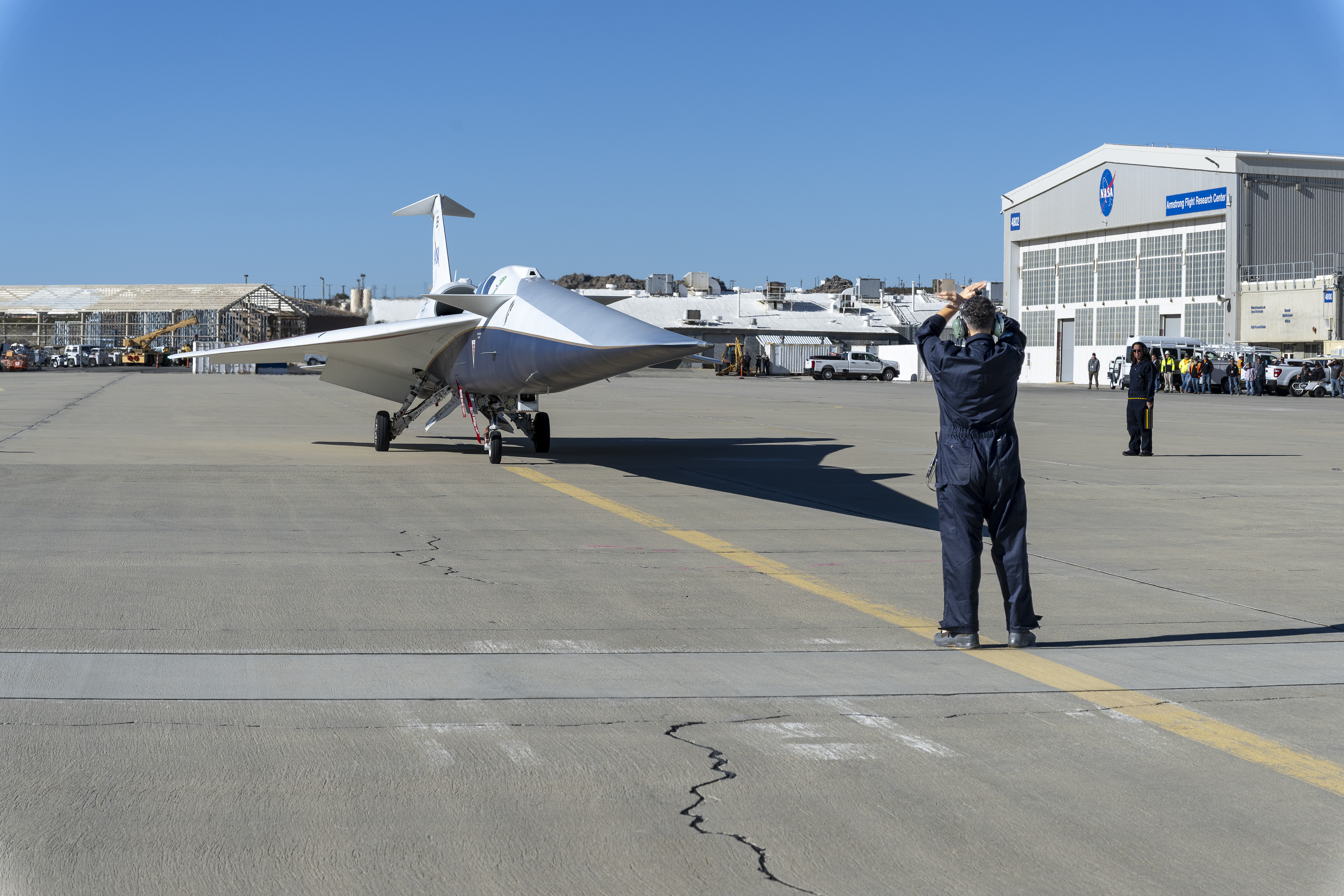 The X-59 pulls onto the ramp at NASA’s Armstrong Flight Research Center. In front of the aircraft, the crew chief stands with arms raised to guide it in, while another flight crew member stands beside the aircraft. An audience is visible in the background near a large hangar.