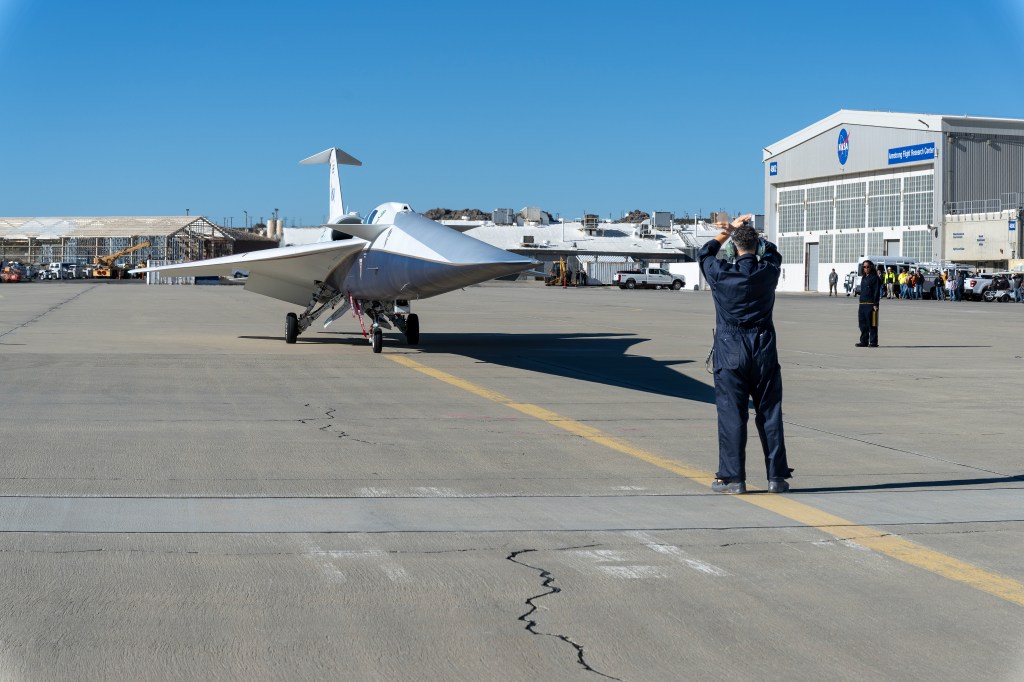 The X-59 pulls onto the ramp at NASA’s Armstrong Flight Research Center. In front of the aircraft, the crew chief stands with arms raised to guide it in, while another flight crew member stands beside the aircraft. An audience is visible in the background near a large hangar.