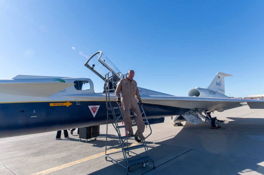 Nils Larson steps down a ladder beside the X-59’s cockpit. He is wearing a tan flight suit with the aircraft parked on the ramp and its canopy open behind him.