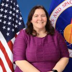 Image shows a photo of Caley Burke mission manager with NASA’s Launch Services Program (LSP) at John F. Kennedy Space Center in Florida posing or a photo with an American flag and NASA flag behind her.