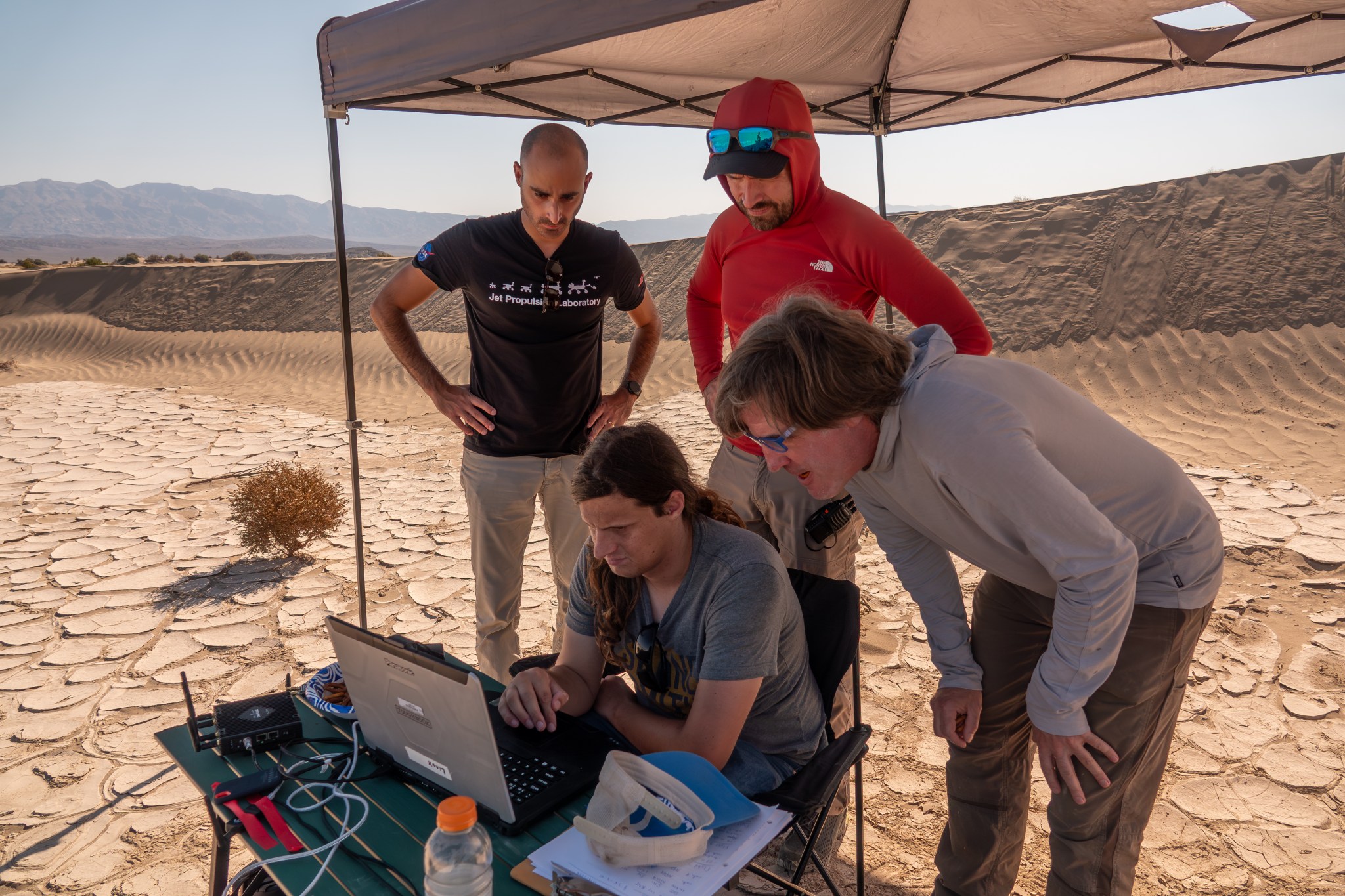 Four people gather around a laptop on a table underneath a tent in the middle of the desert.