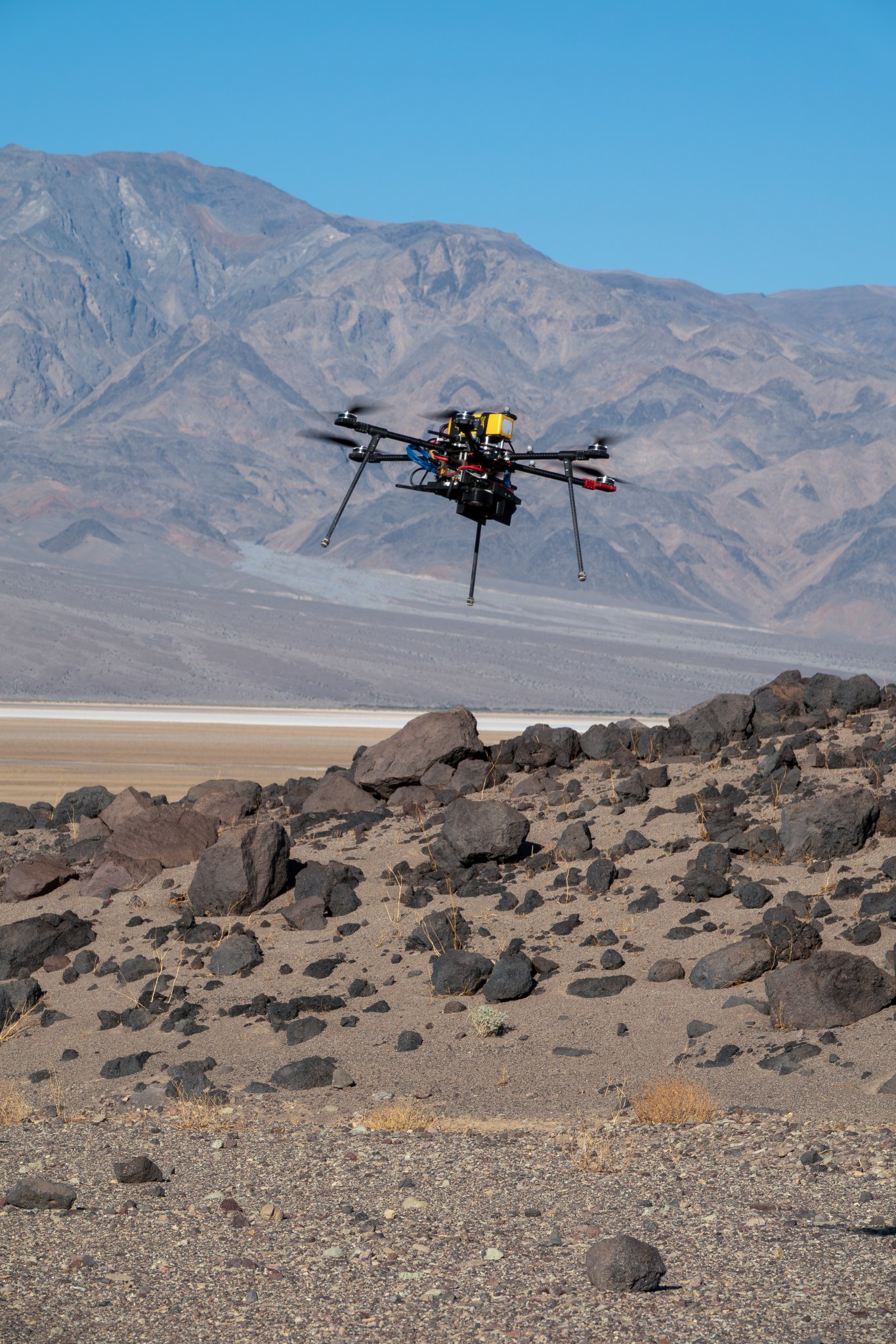 A drone flies over a rocky desert surface with a mountain and blue sky in the background.