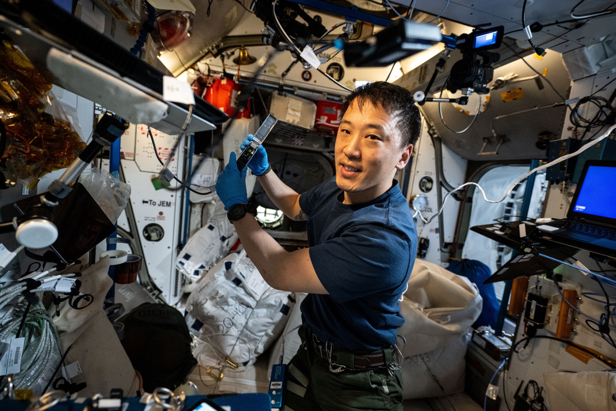 NASA astronaut Jonny Kim, wearing a navy-blue shirt and blue latex gloves, holds up a rectangular, controller-sized device with both hands. He is surrounded by electronic equipment and wires.