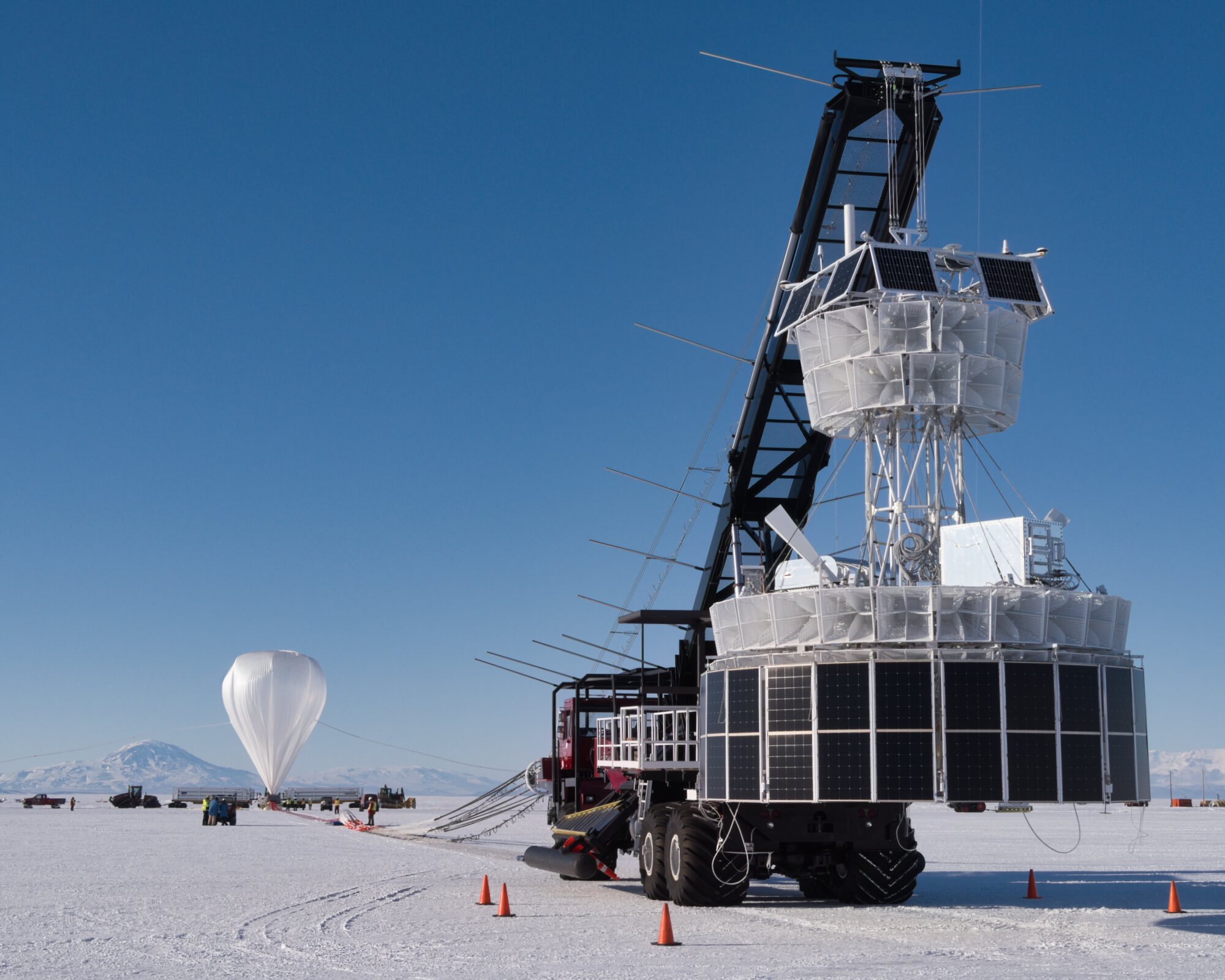 A large metal scientific instrument that is mainly white with black panels is suspended off the ground by a large crane. A white trail of material is on the ground and attaches to a partially inflated white balloon in the background on a snowy landscape.