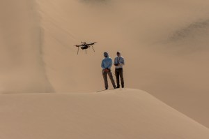 Two people stand atop a sand dune, facing the camera. They are both looking at a drone flying in front of them. The person on the right holds a controller. Other sand dunes behind them make up the background of the entire image.