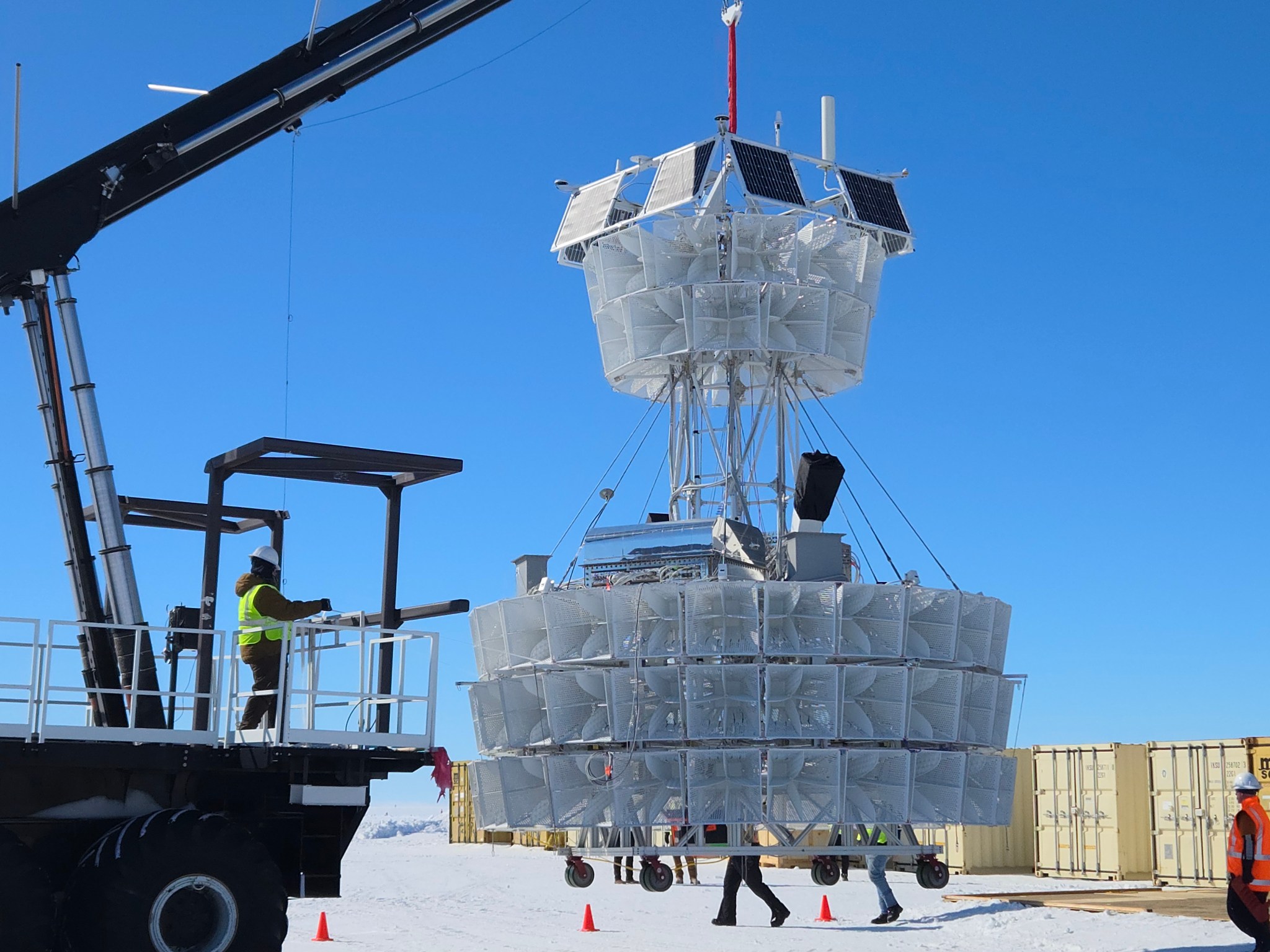 A large scientific balloon payload is being lifted by a black crane against a bright blue Antarctic sky. The payload consists of multiple tiers of white protective panels or covers arranged in a cylindrical configuration, with solar panels visible on the top section and various antennas protruding upward. The equipment is suspended by cables and positioned over a wheeled transport cart on the snow-covered ground. A technician in a bright yellow safety vest operates the crane from an elevated platform on the left side of the image