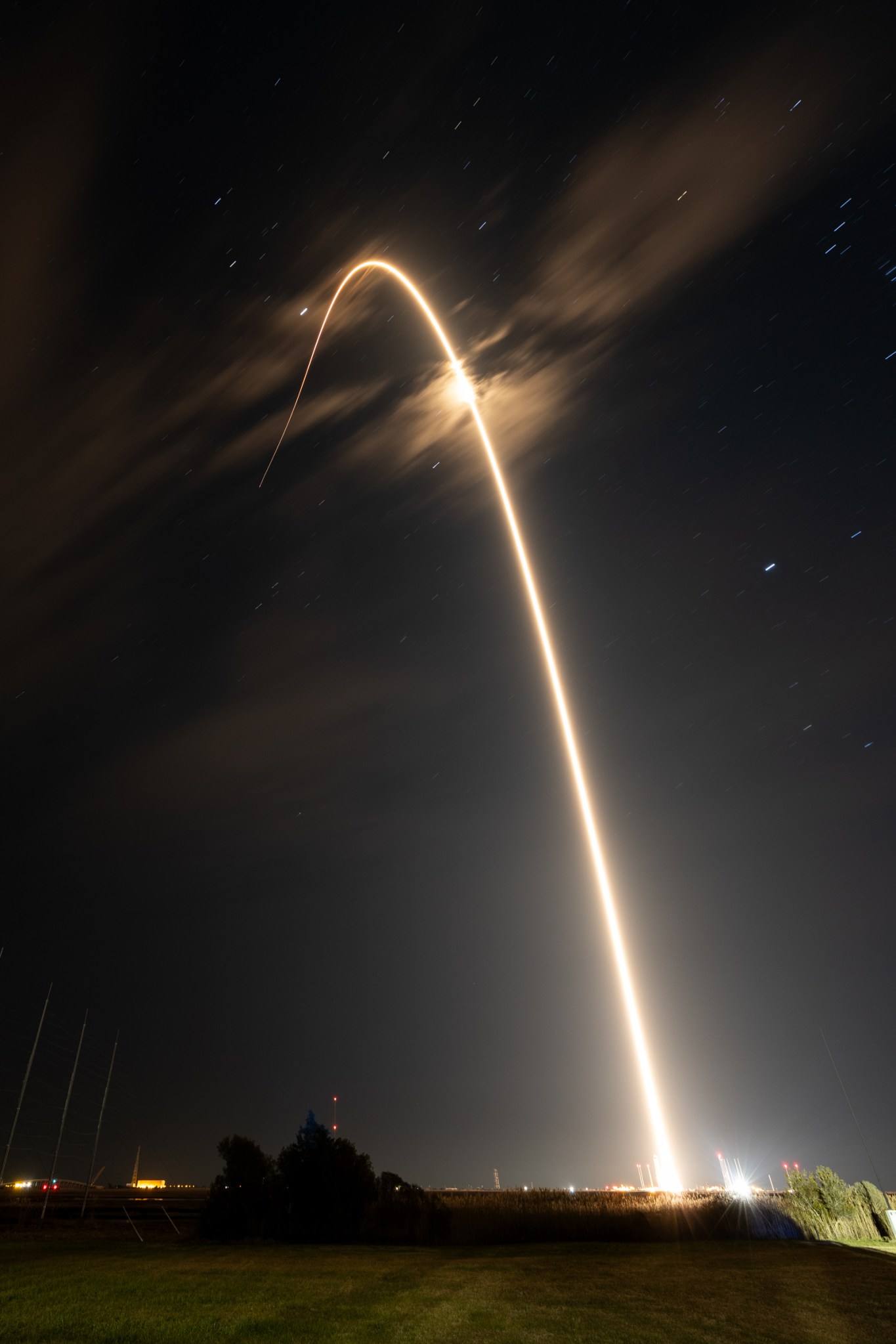 A long exposure photograph of the Electron launch, which is a white streak through a small patch of clouds across a black sky with stars.