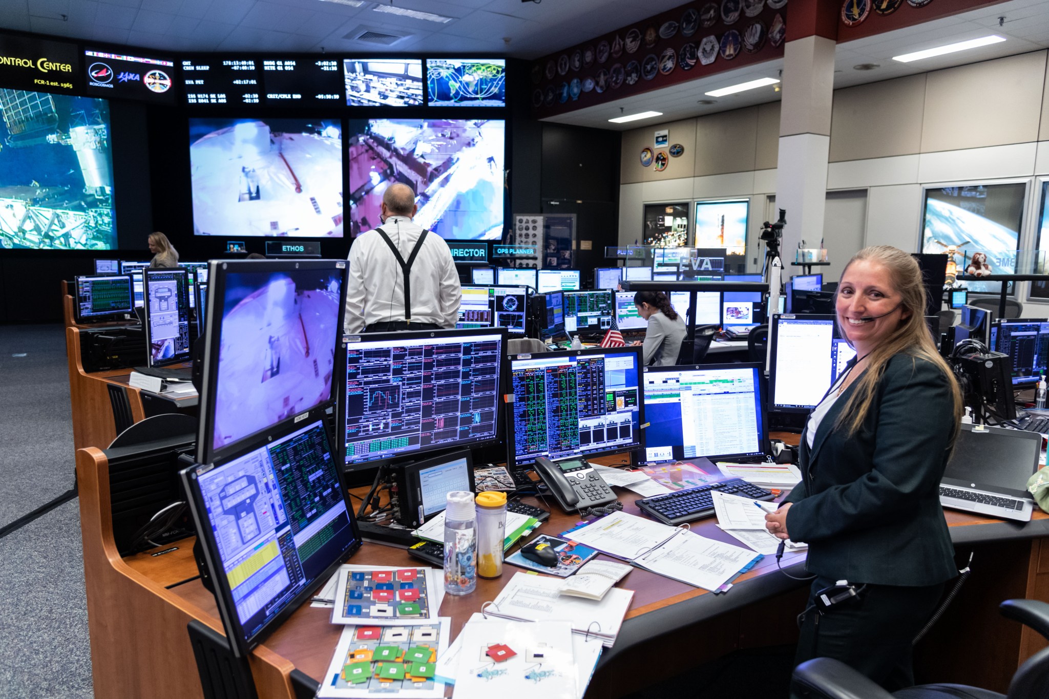 A woman smiles and poses at a desk in front of several monitors at mission control.