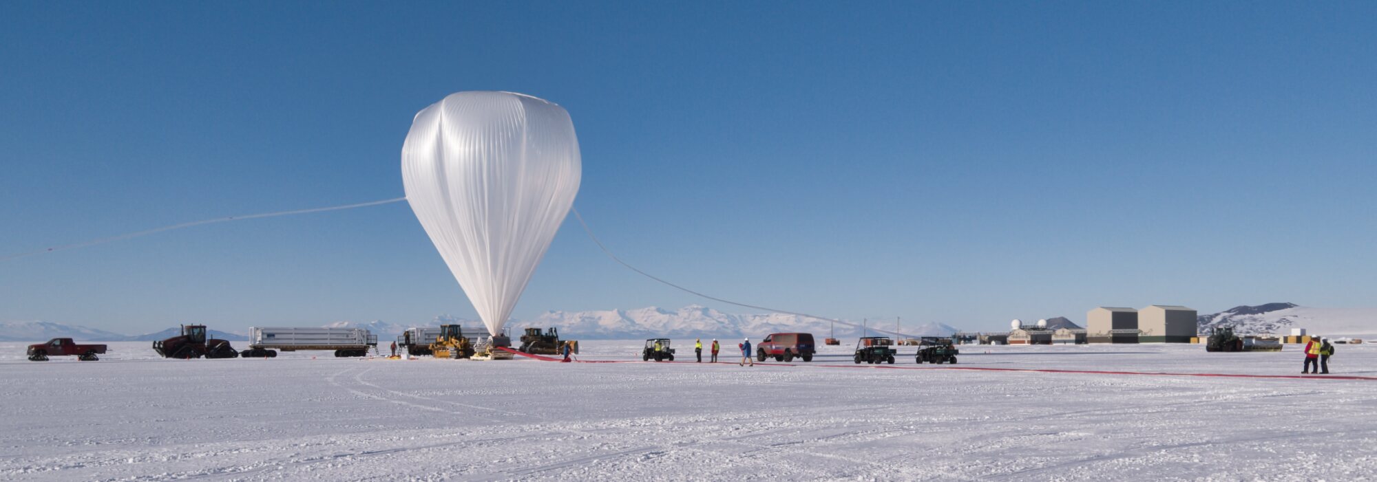 A fully inflated white scientific balloon tethered on the Antarctic ice with ground crew, support vehicles, and facility buildings visible across the snow-covered landscape under a clear blue sky