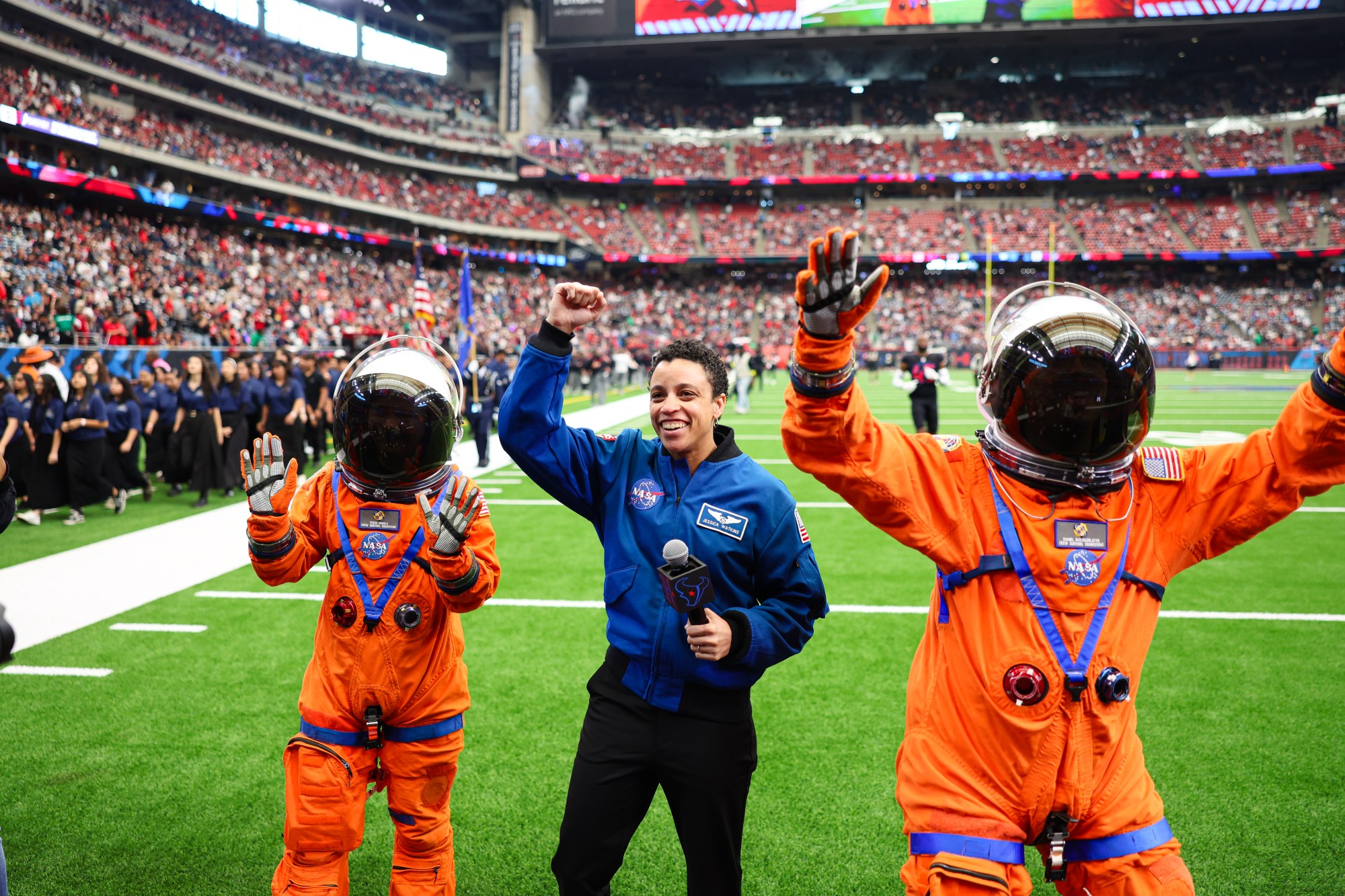 Three people pose with their hands in the air on the football field. Two people are wearing an orange spacesuit and the person in the middle is wearing a blue flight jacket.