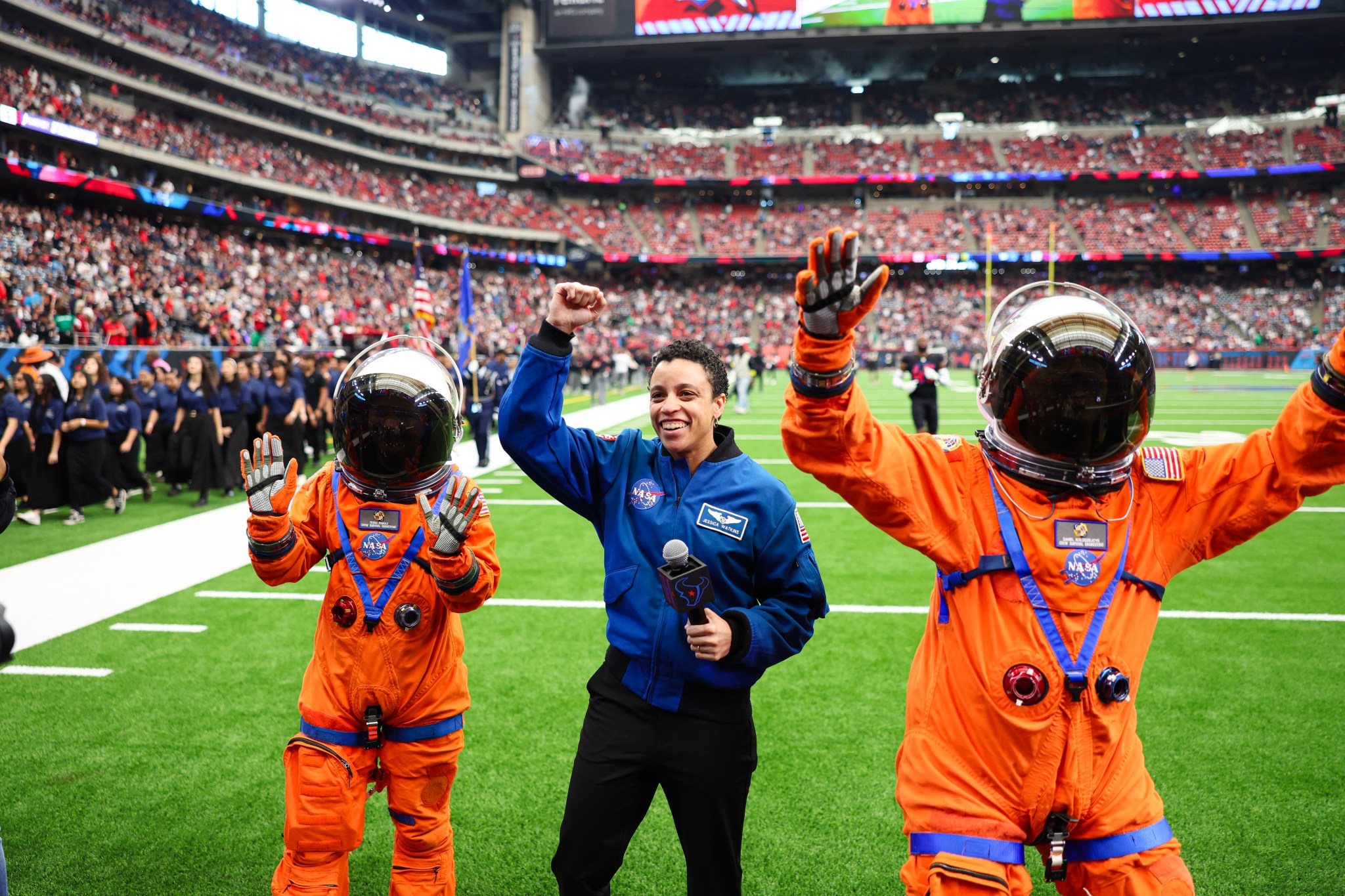 Three people pose with their hands in the air on the football field. Two people are wearing an orange spacesuit and the person in the middle is wearing a blue flight jacket.