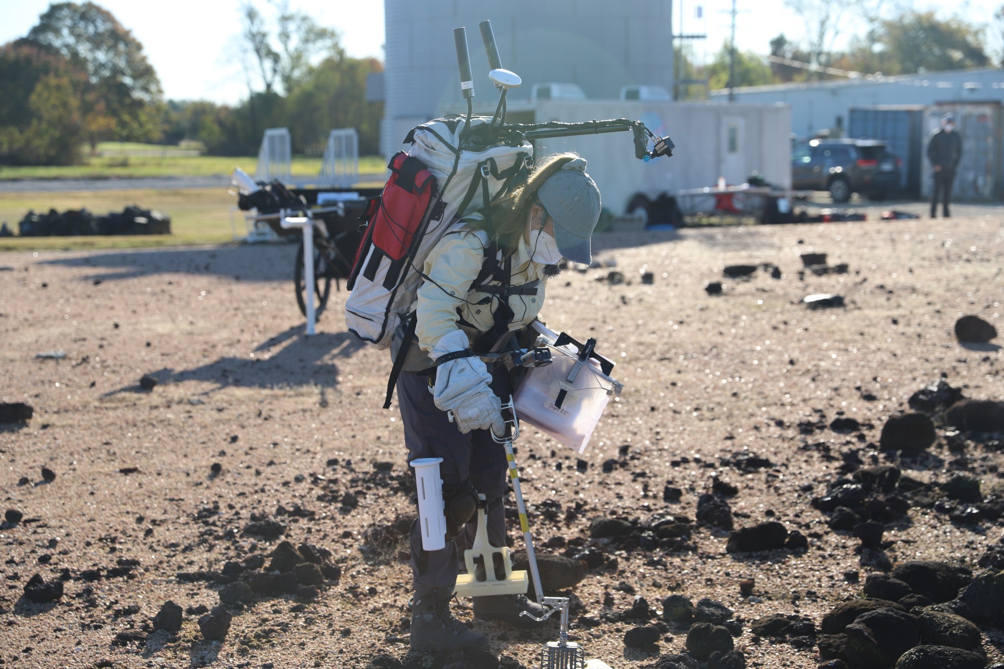 A woman wears a lunar backpack while practicing picking up rocks with a lunar tool at a rock yard.