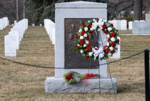 A wreath with red and white flowers, green leaves, and white ribbon bows rests on a tripod stand with three thin metal legs. It stands in front of the Space Shuttle Challenger Memorial. The Memorial has two parts: A polished gray stone headstone with a bronze embossed plaque on it, resting on a small platform of rough gray stone. The plaque has the likeness of Challenger and the crew on it. Behind the memorial, we can see rows of white stone headstones.