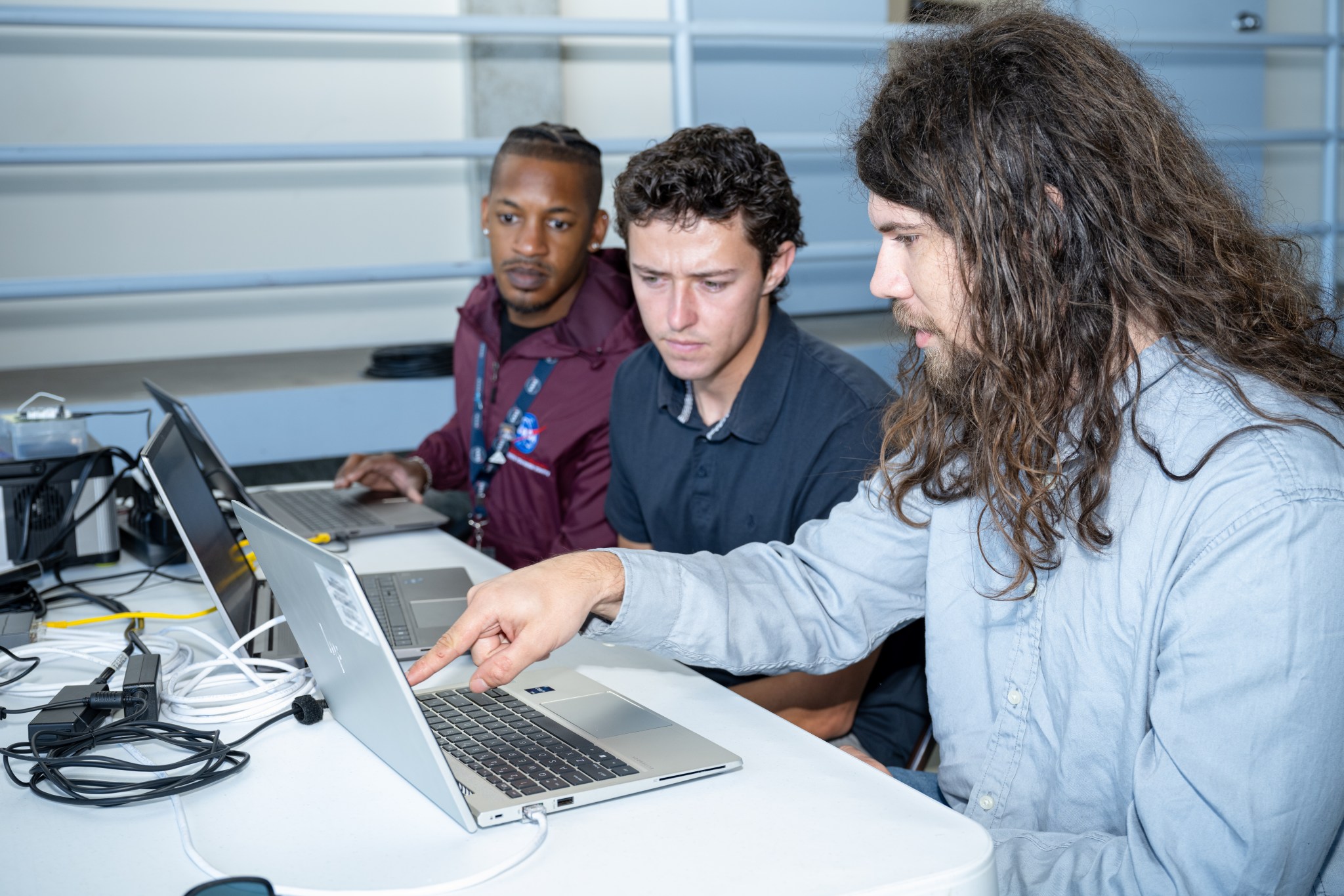 Three people sit at a table looking over laptop computers. The one nearest the camera is pointing at the laptop monitor.