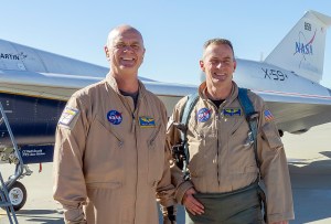 Two NASA pilots wearing flight gear pose in front of the X-59 experimental aircraft.