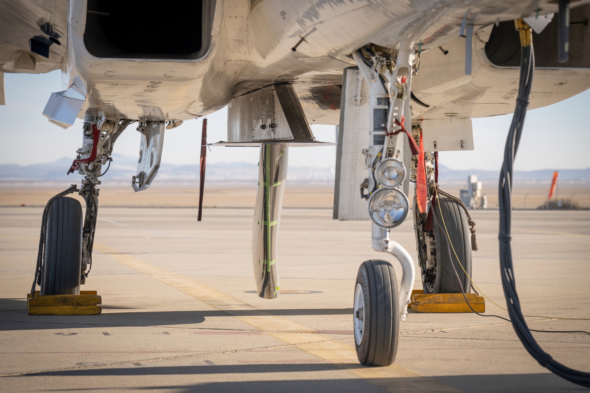 A NASA F-15 research aircraft is parked on a ramp at NASA’s Armstrong Flight Research Center in Edwards, California, with an experimental wing design mounted beneath its fuselage. The gray and silver test article is positioned vertically, resembling a ventral fin.
