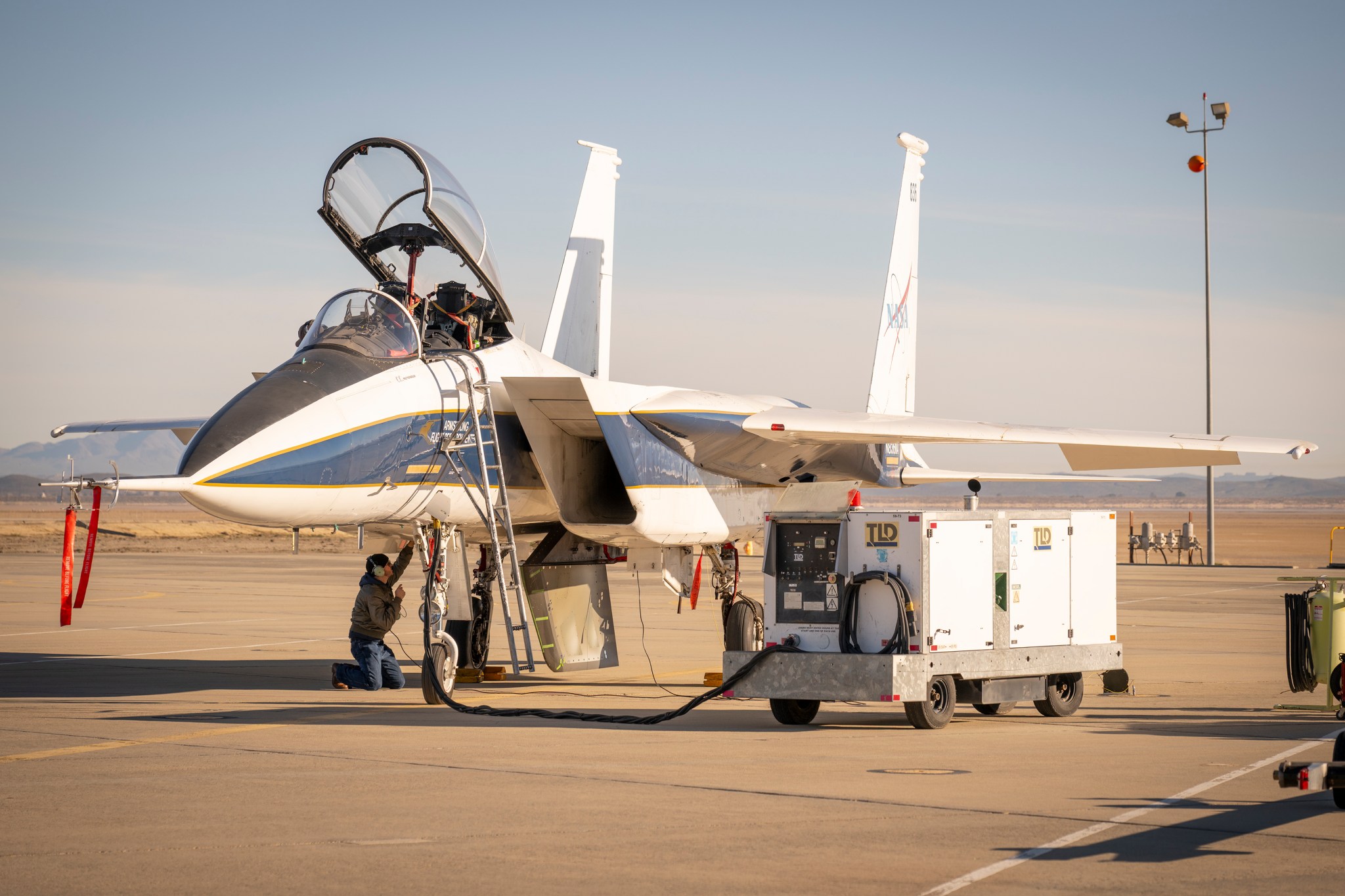 A NASA F-15 research aircraft is parked on a ramp at NASA’s Armstrong Flight Research Center in Edwards, California. Ground crew work beneath the aircraft on an experimental test article, resembling a ventral fin, mounted under the aircraft’s fuselage.