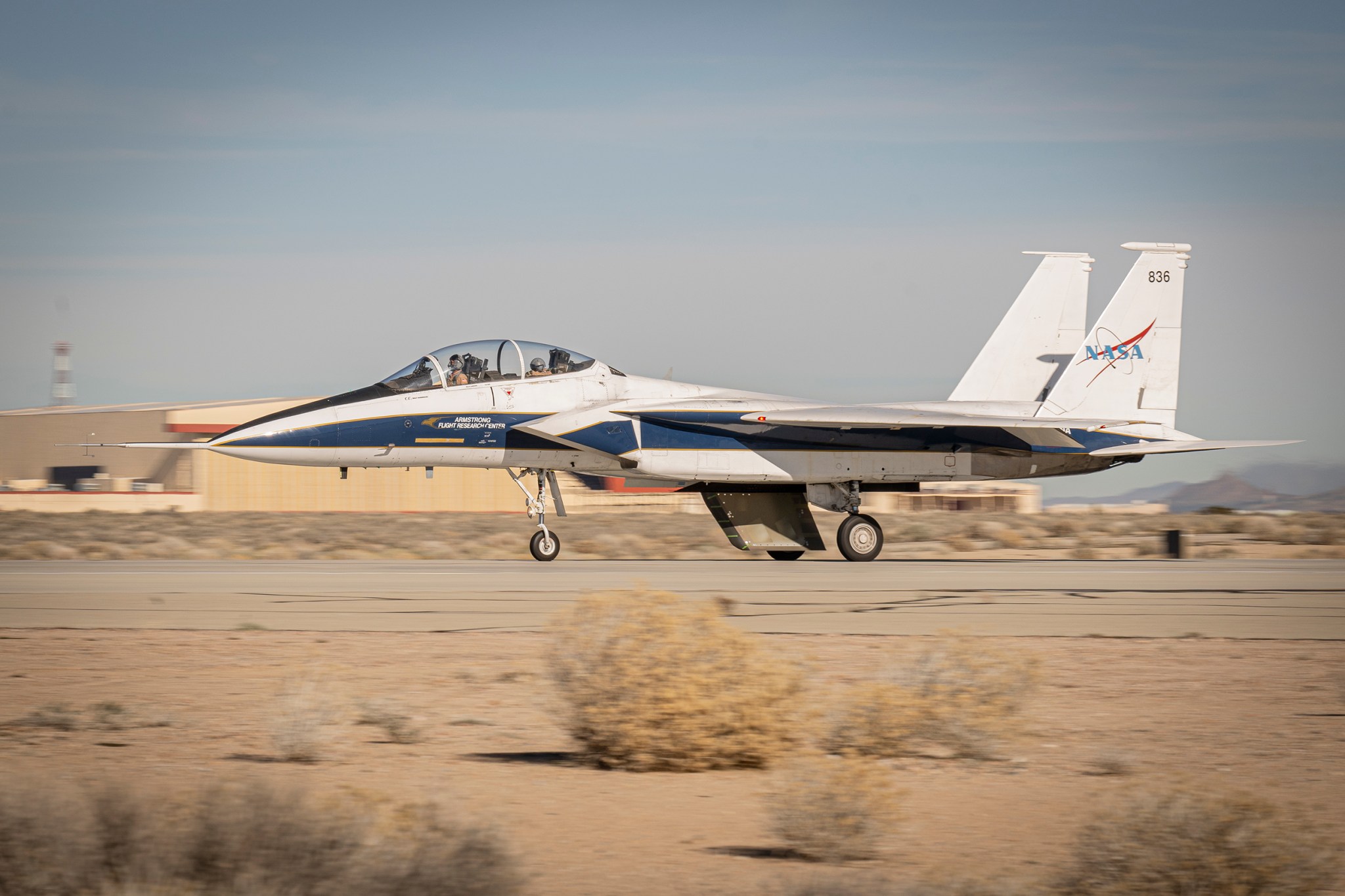 A white and blue NASA F-15 research aircraft taxis down a runway at Edwards Air Force Base with an experimental wing design mounted beneath the fuselage, resembling a ventral fin. In the background, a desert landscape with mountains and tan buildings stretches as the aircraft moves past.