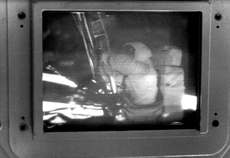 Black-and-white image displayed on a CRT monitor showing a grainy television transmission of Armstrong from the waist-up climbing down to the lunar surface, with parts of a ladder and spacecraft structure visible nearby. The image is framed by the monitor’s metal casing, emphasizing the low-resolution, historic broadcast quality.