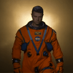 Artemis II Commander Reid Wiseman, wearing an orange Orion Crew Survival System spacesuit, steps forward in front of a golden light.
