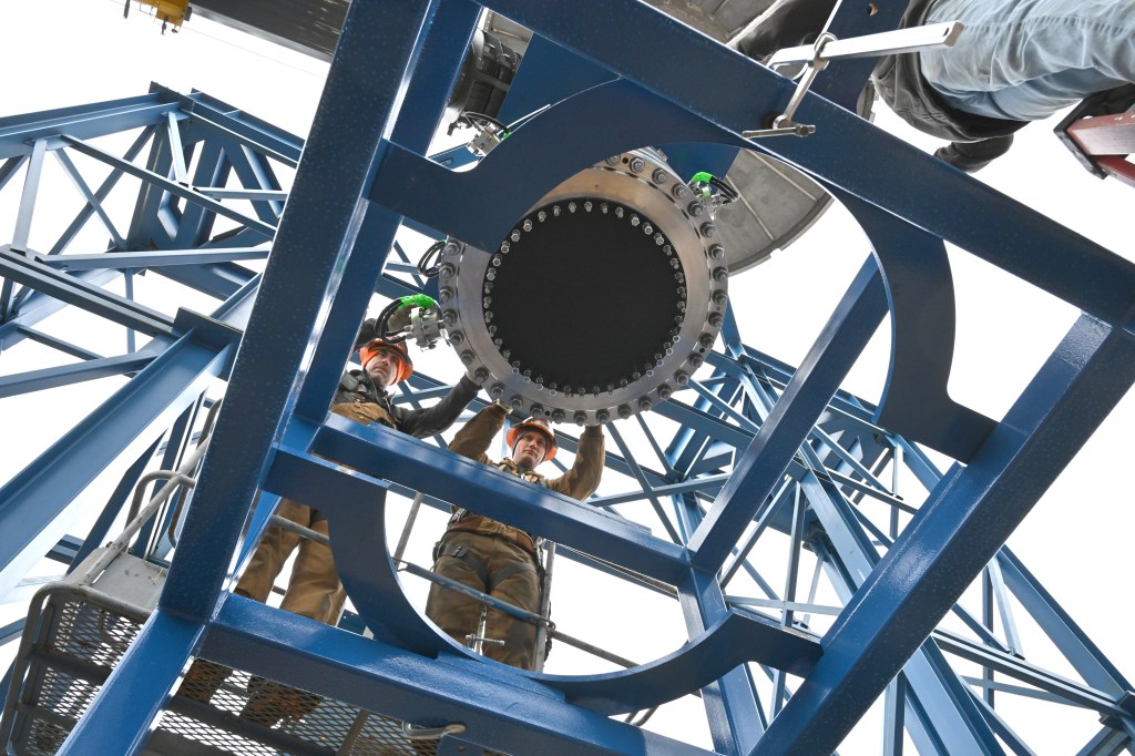 Overhead view looking up through a blue steel test stand structure at a large cylindrical rocket motor with a circular black nozzle opening and bolt-pattern flange. Multiple workers in safety gear are positioned around the hardware on the platform.