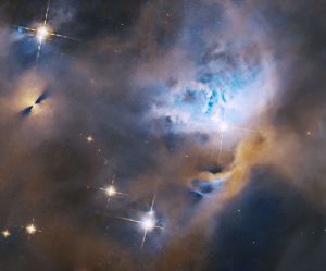 Within a ghostly blue and orange cloud of gas and dust, several stars shine brightly. To the upper left is a protoplanetary disk, appearing as a bright point flanked on either side by fan-shaped shadows. To the center right, stars are nestled in a cavity in the gas and dust.
