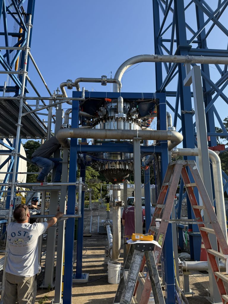 A rocket engine with dual nozzles mounted on a blue test stand surrounded by stainless steel propellant feed lines and piping. A technician in a white shirt observes from the lower left while ladders and support equipment are positioned around the test article. Clear blue sky and trees are visible in the background.