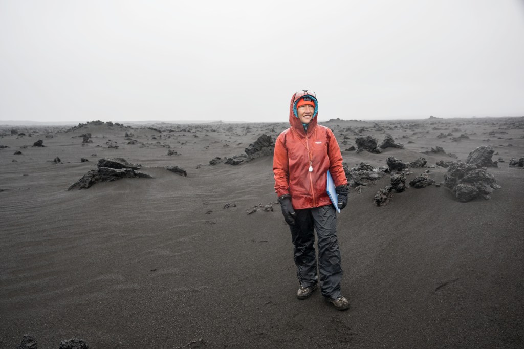 Christina Koch stands in a red rain jacket on a sandy grey Moon-like landscape in Iceland.