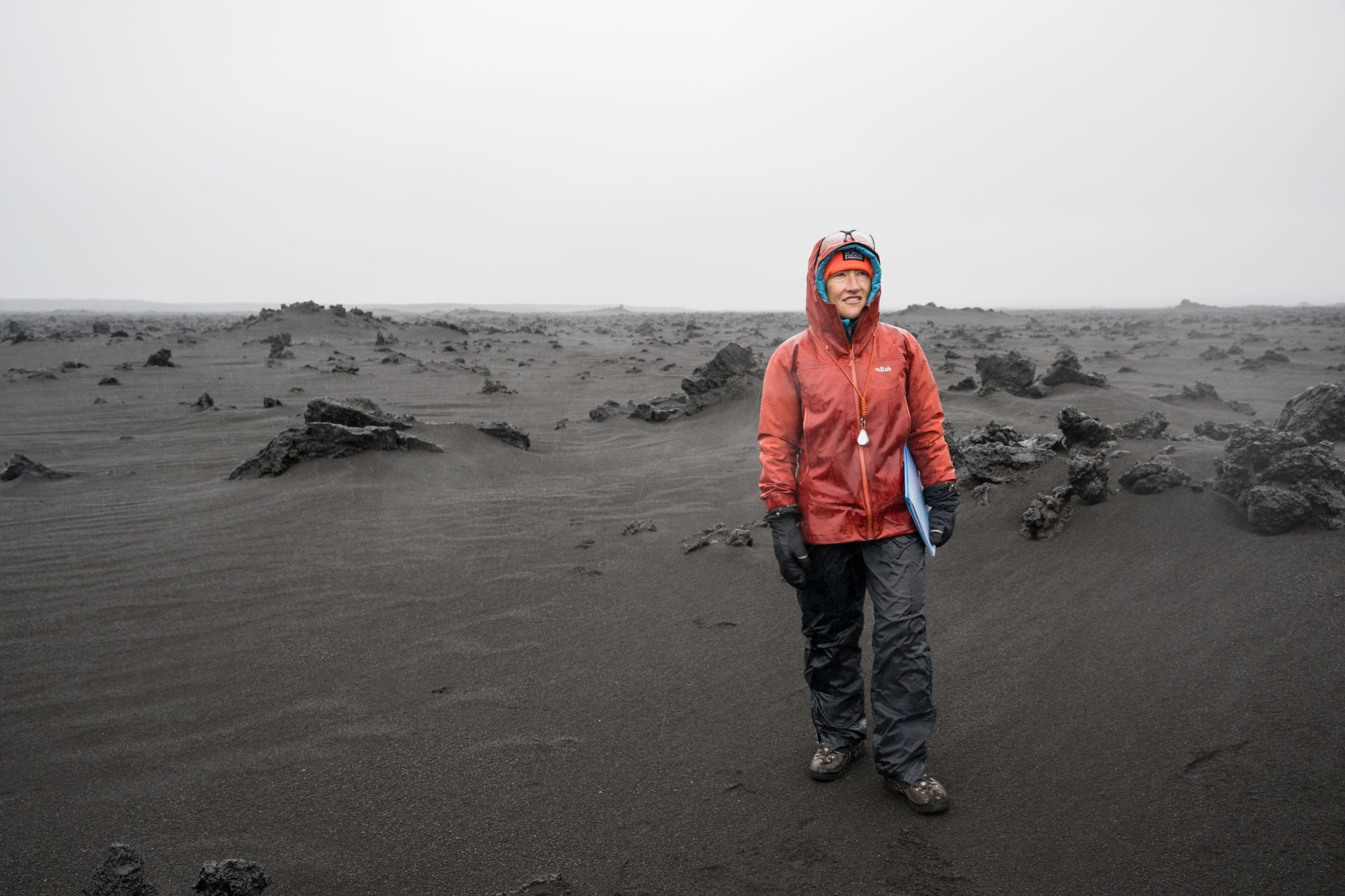 A woman wearing an orange jacket holding a folder poses in a rocky terrain.