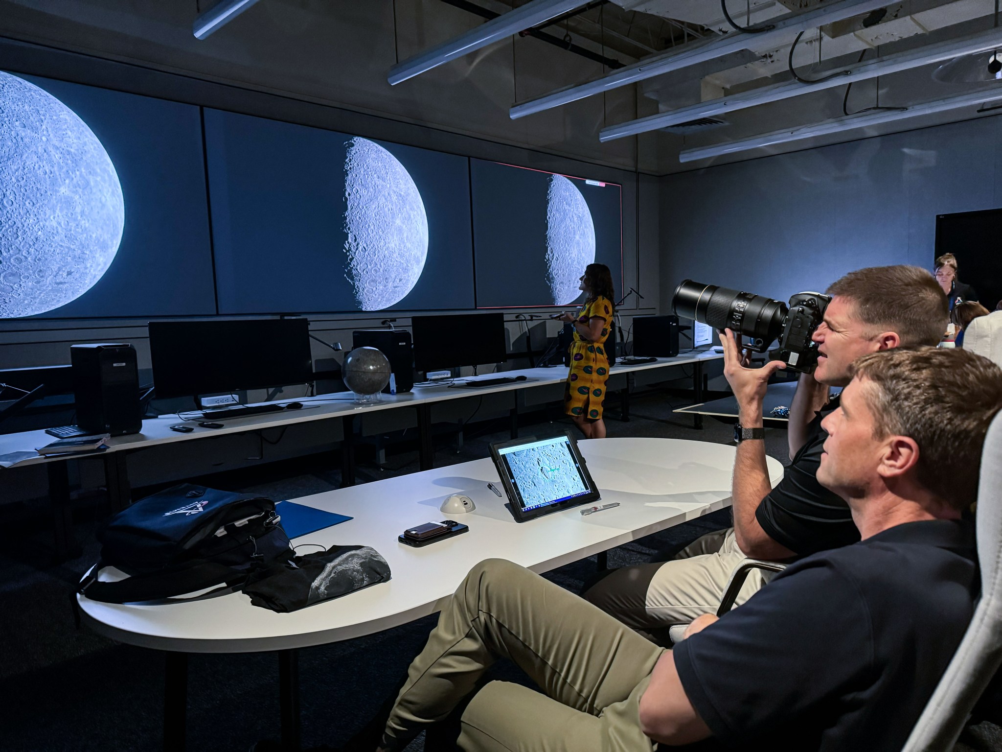 Two people observe phases of the moon inside of a room. The person on the right is holding a camera.