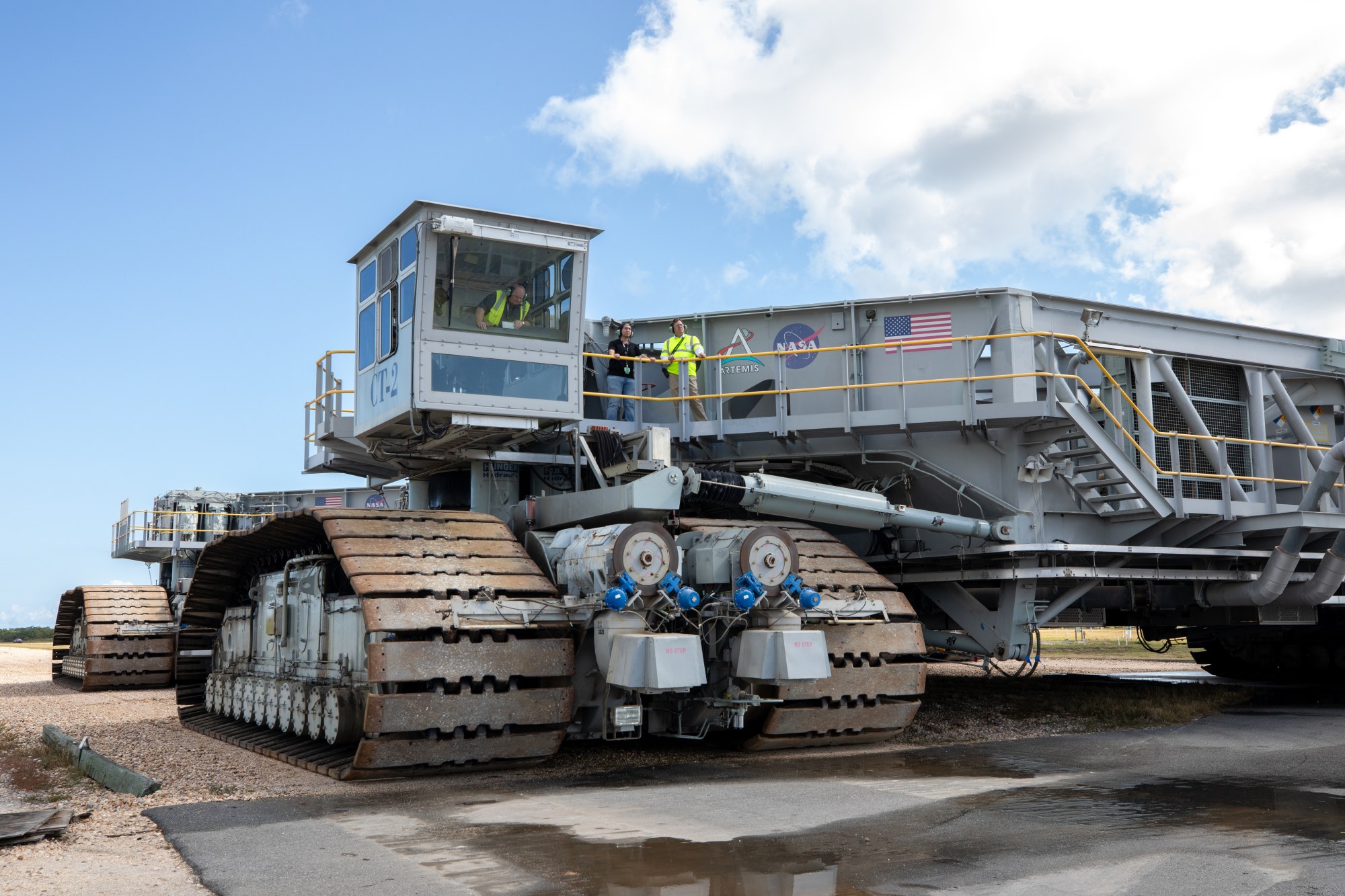 A crawler-transporter, including its massive treads take up the majority of the image. In fact, the entire crawler cannot be seen. There are a few people at the top of the crawler, which allows you to see just how massive the machine is.