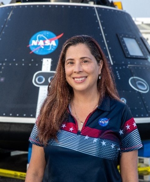 A woman with brown hair smiles for the camera while standing in front of the NASA Artemis Orion spacecraft. The spacecraft with a NASA insignia displayed is cone shaped and black and silver in color.