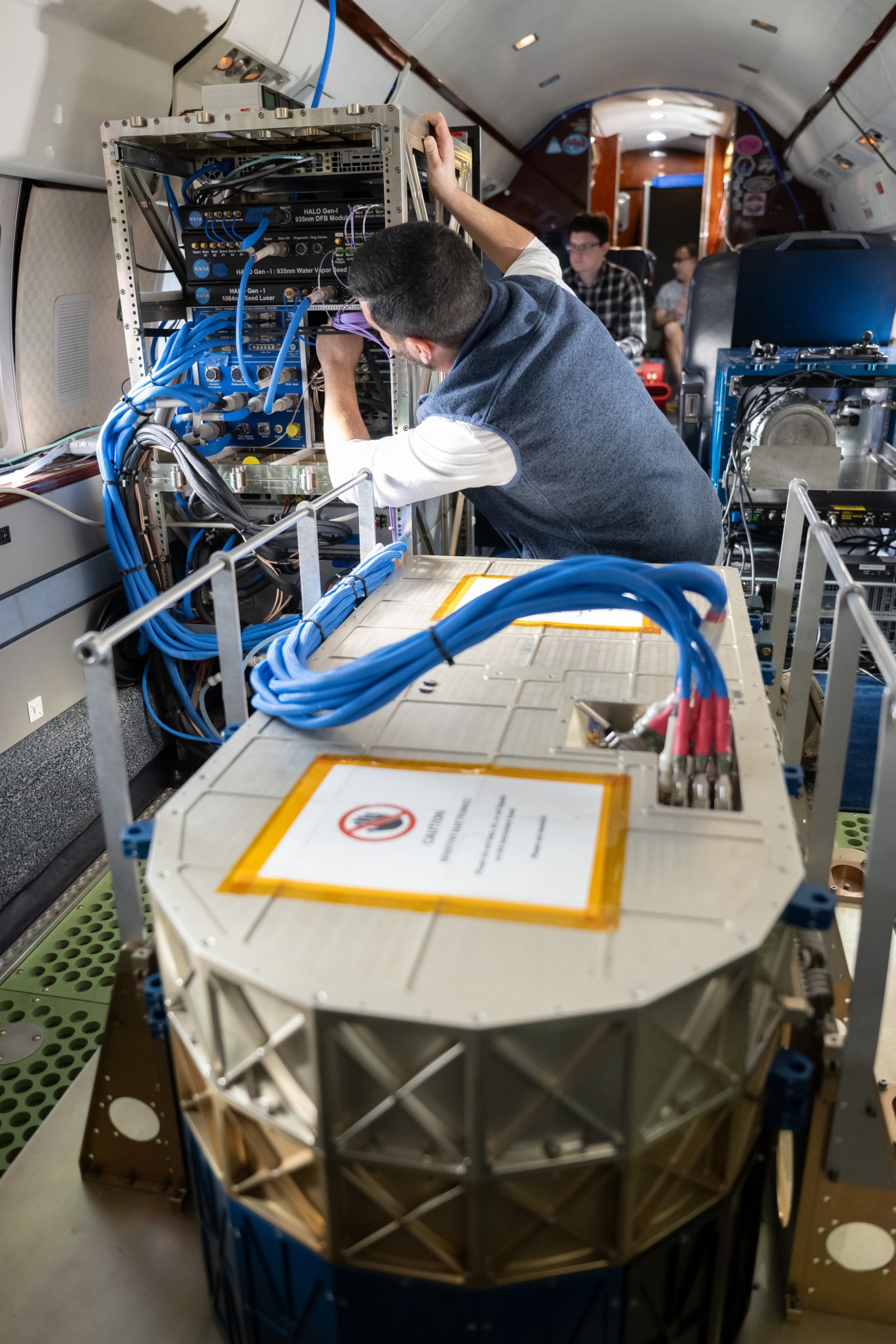 An oval shaped, metal instrument about one foot tall and four feet long is on the foreground floor inside a small aircraft cabin. A researcher can be seen connecting cables from the instrument to a rack of computer drives.