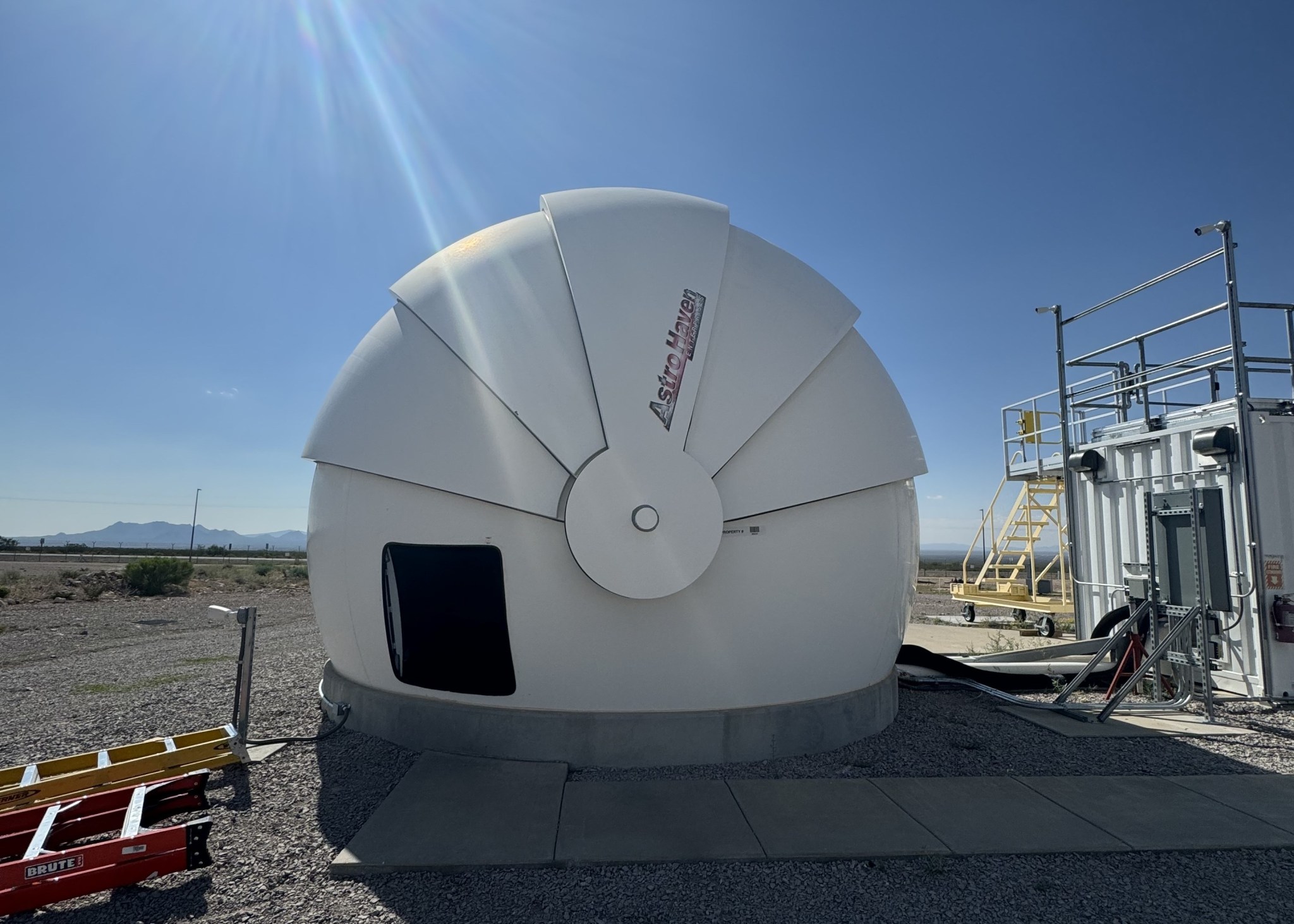 A white ground station is pictured against a bright blue sky with the sun in the upper lefthand corner. The ground station looks like a snail's shell and casts a shadow off toward the right.