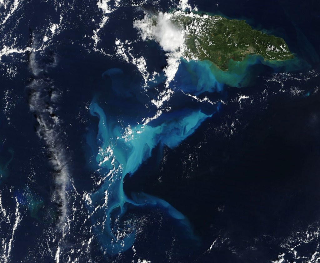 A satellite image shows a portion of the dark blue Caribbean Sea near Jamaica. A submerged carbonate platform appears as a slightly brighter blue area of water in the center. The mostly green island of Jamaica is in the upper right, and scattered clouds are present throughout.