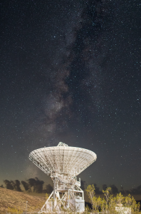 A white radio antenna faces upward in the direction of a star-studded night sky.