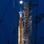 NASA's Space Launch System rocket, which is orange and white, stands on the launch pad with the mobile launcher platform. A full Moon hangs in the sky.
