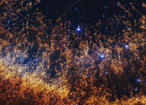 A closeup of a small section of the Helix Nebula, an expanding shell of gas and dust. Thousands of orange and gold comet-like pillars stream upward from the bottom, like thin liquid blown up a sheet of glass. These pillars are around the circumference of the arced shell, which forms a partial orange semi-circle at the bottom. The pillars are more numerous and denser at the bottom, and darker red. They fade to orange and then yellow in the arc. In the top two-thirds, they are thinner and more golden, and it’s easier to see the black background of space. Several bright blue stars, some with diffraction spikes, are scattered throughout. A few larger stars are on the right side.