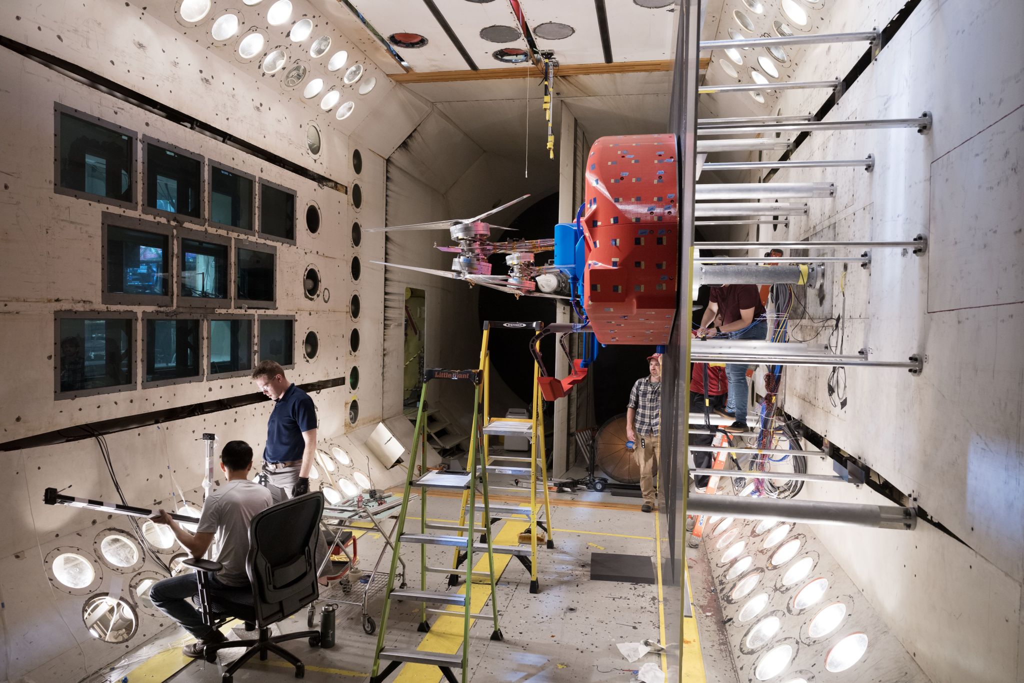 Five people in a large white room work in and around a red car-sized rotorcraft prototype mounted on a wall.