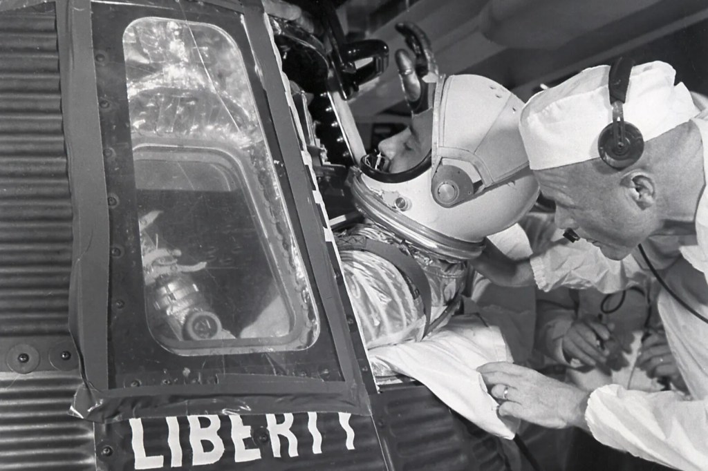 Black-and-white photograph showing an astronaut being seated inside the Liberty Bell 7 Mercury spacecraft during preflight preparations, while two technicians in white caps and headsets assist at the open hatch. The capsule’s exterior is visible in the foreground with the hand-painted text “LIBERTY BELL 7” on its corrugated metal surface.