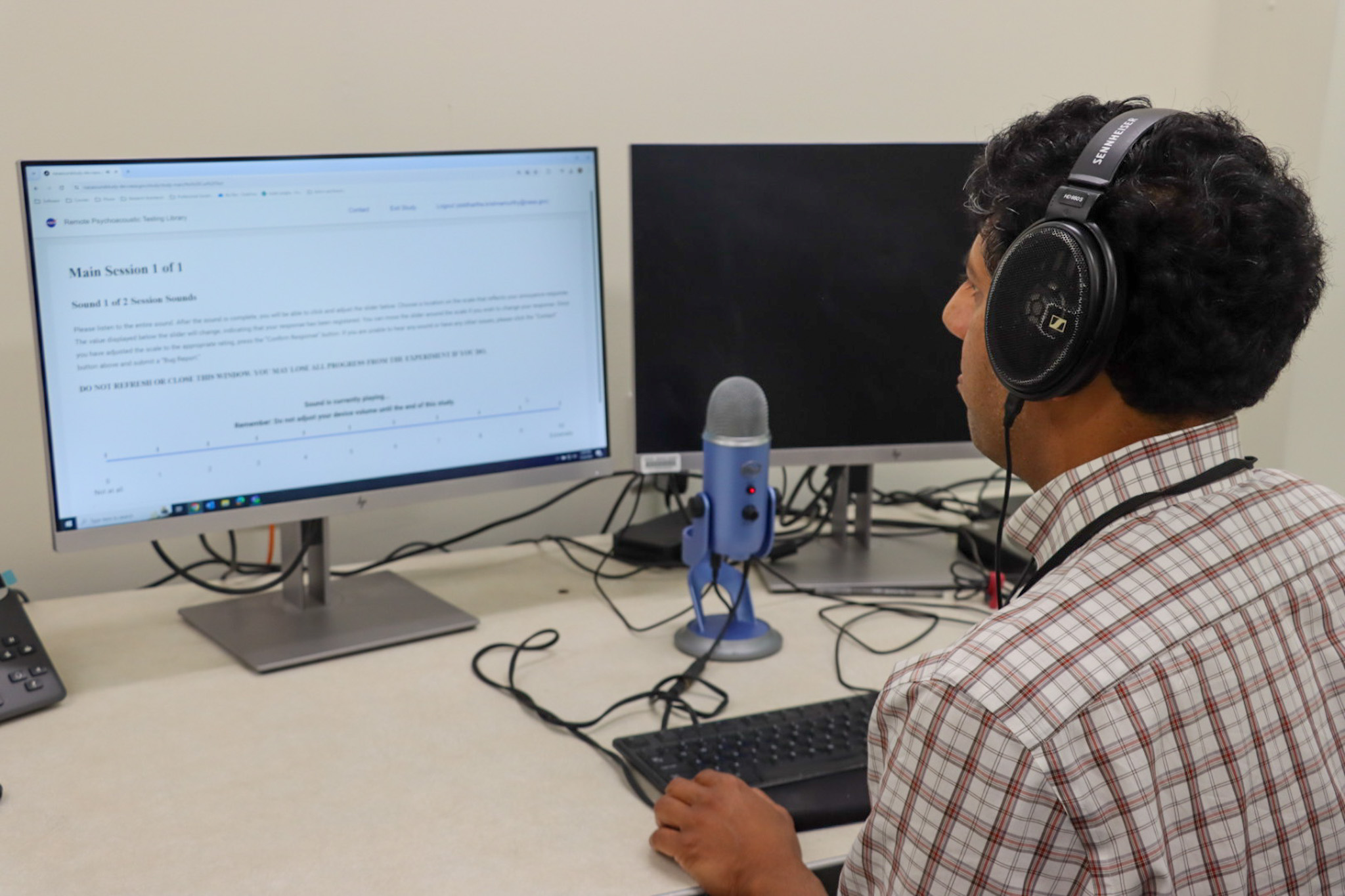 A man wearing a white and red plaid shirt sits to the right side of the image in front of a keyboard, a blue stand microphone, and two monitors. He wears black over ear headphones and looks at a computer screen with a white background and black letters.