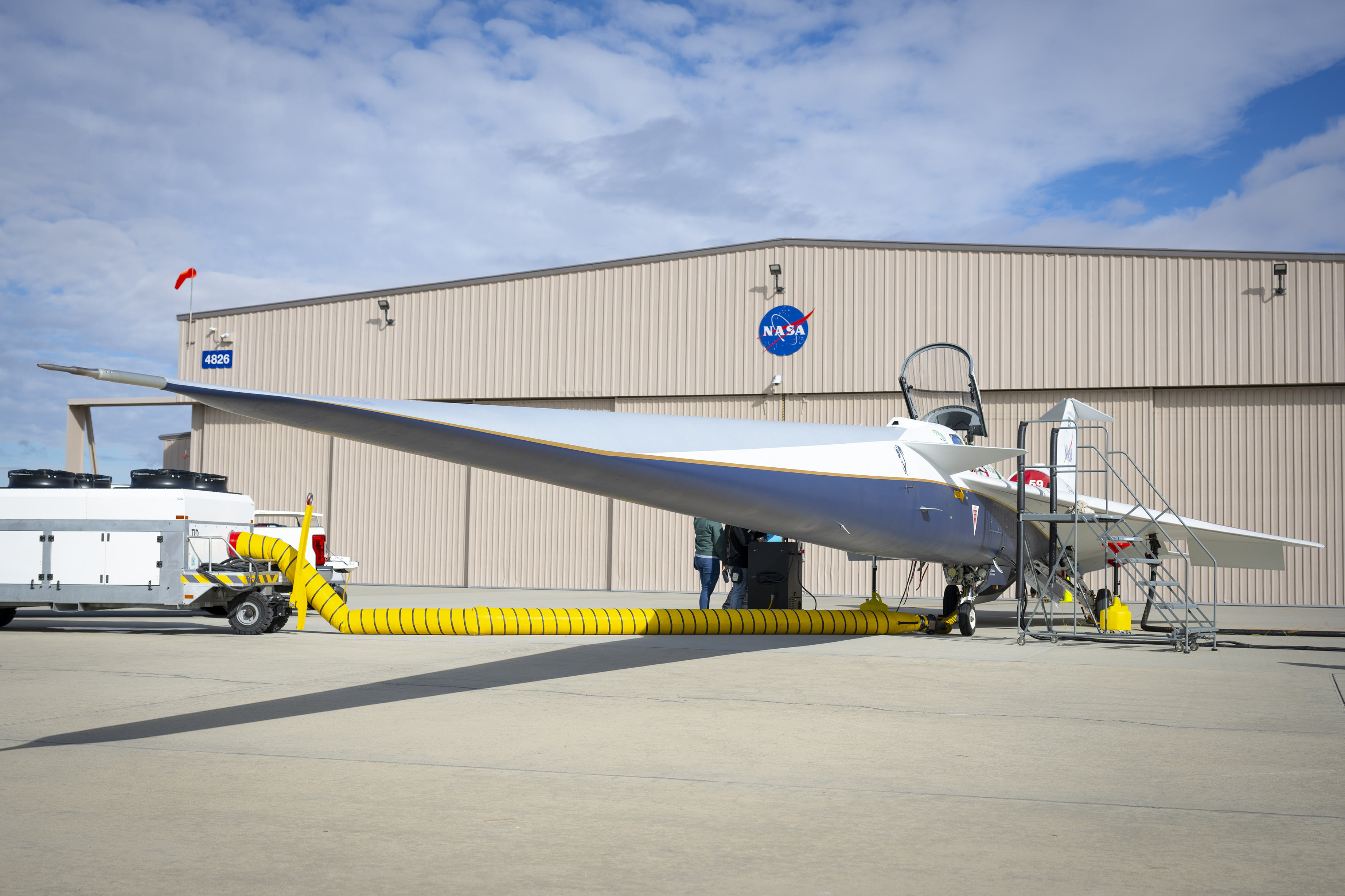 A white and blue jet airplane is parked in front of a building with large sliding doors and a NASA logo centered on the forward wall. The building is the new X-59 hangar.