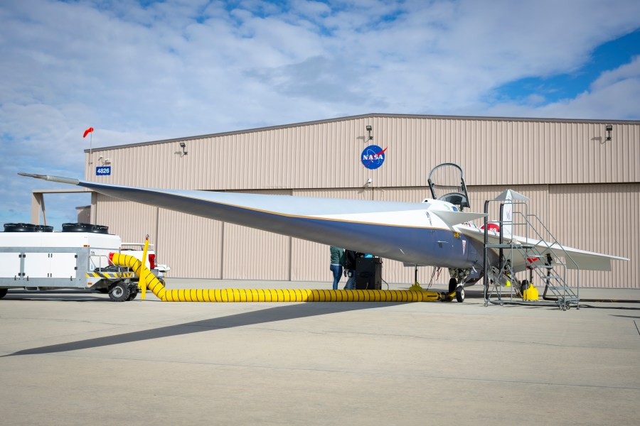 A white and blue jet airplane is parked in front of a building with large sliding doors and a NASA logo centered on the forward wall. The building is the new X-59 hangar.