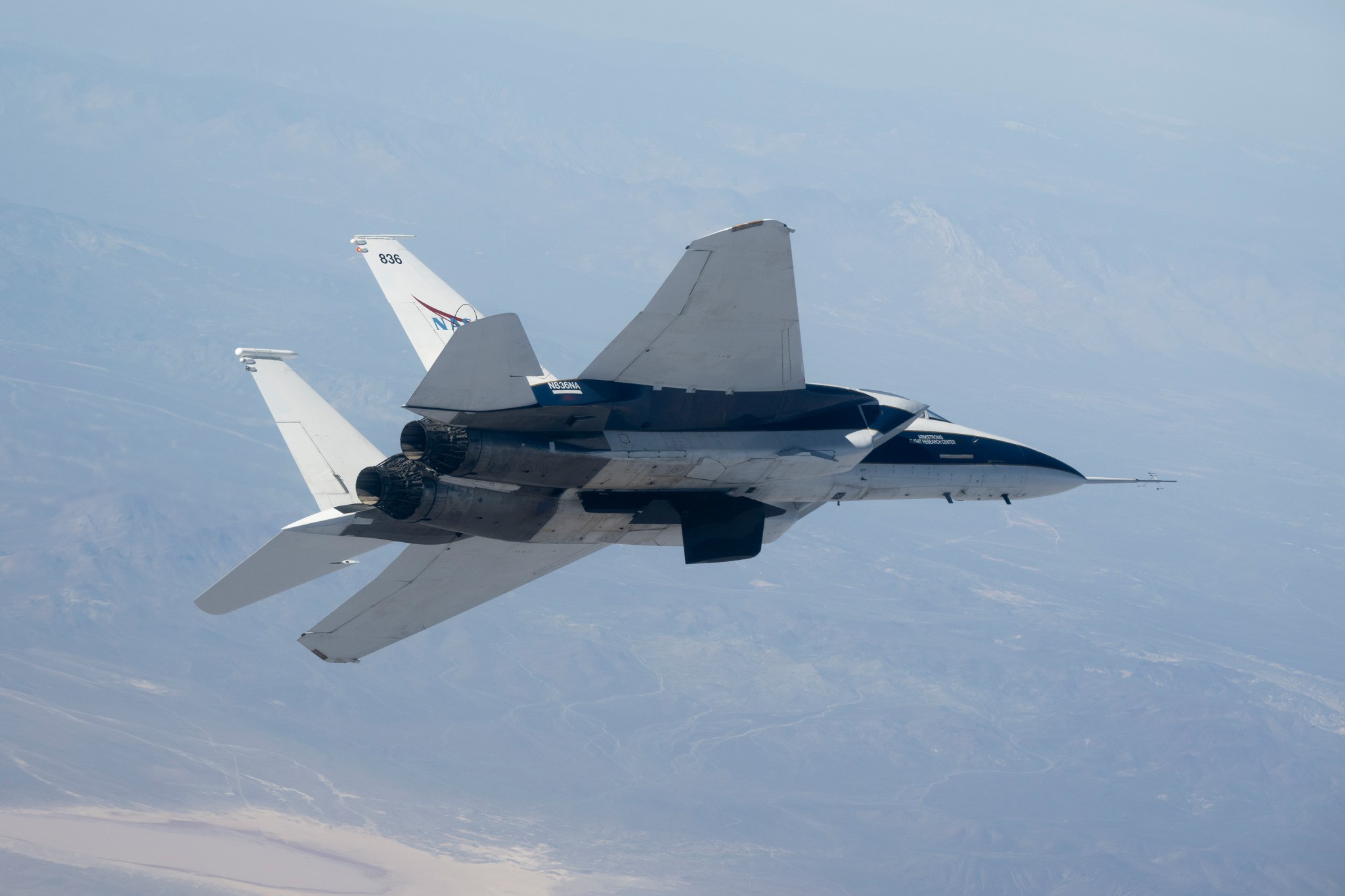 A NASA F-15 research jet flies over the California desert with an experimental wing design attached beneath its fuselage, shown in profile above a dry lakebed and a nearby city during the first flight of the experimental wing.