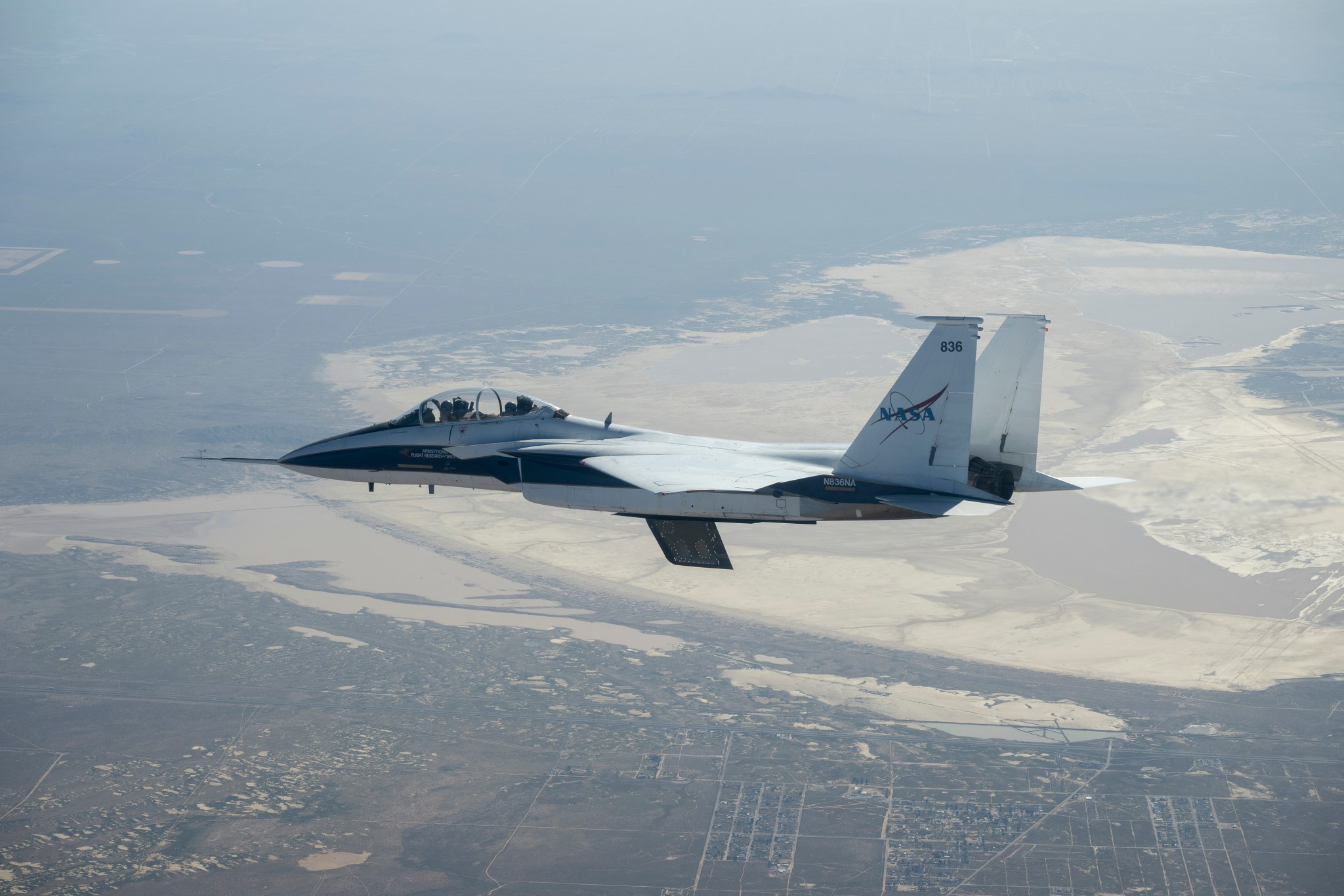 A white and blue NASA F-15 research jet climbs to altitude with an approximately 3-foot experimental wing design mounted beneath its fuselage. Viewed in profile against a blue sky with mountains in the distance, the test article resembles a ventral fin below the aircraft.